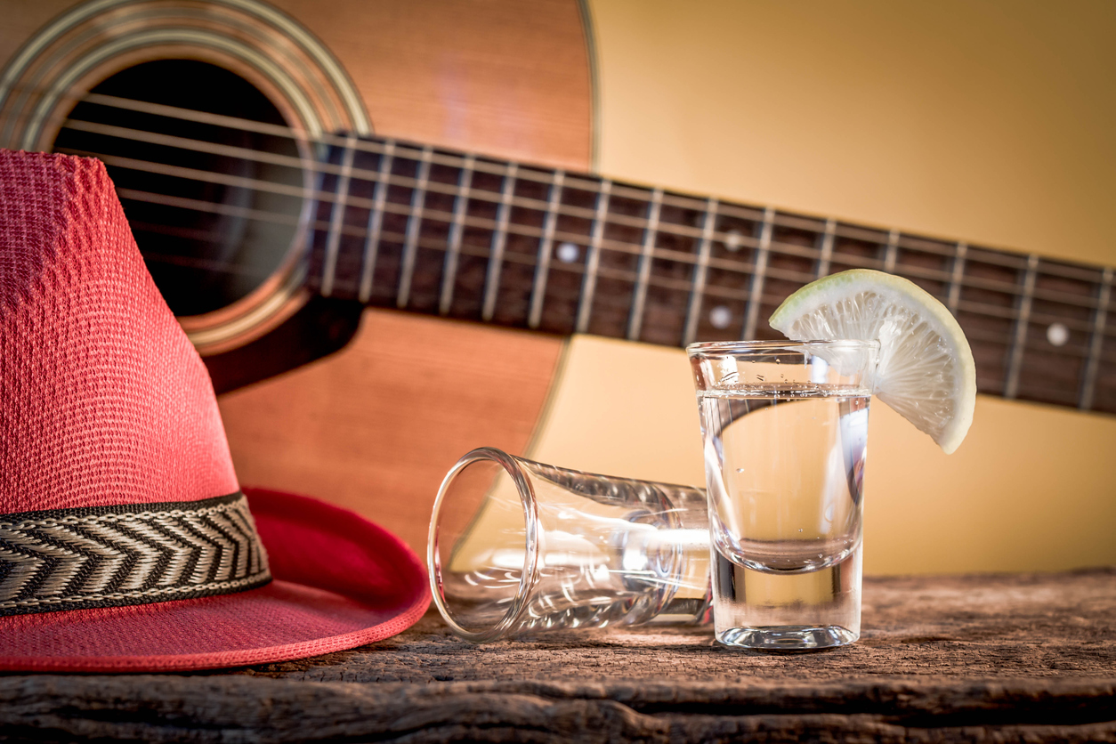 A glass of clear liquor sits beside a classical guitar topped with a red hat, creating a festive and vintage vibe—perfect inspiration for a Tequila Playlist theme.